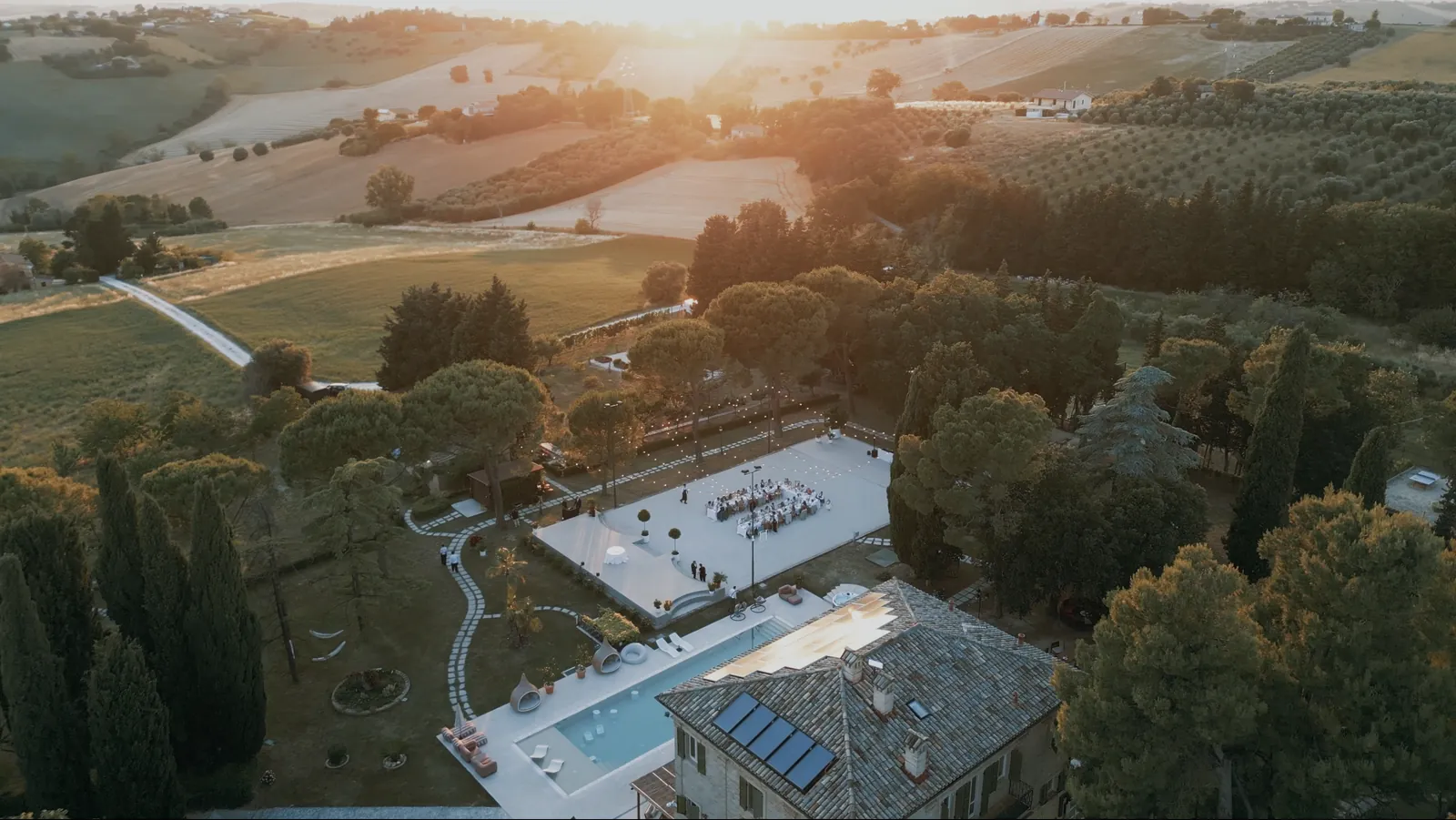 Drone view of Maniero Montecassiano at sunset, with dinner set on the lawn court above the pool and the villa