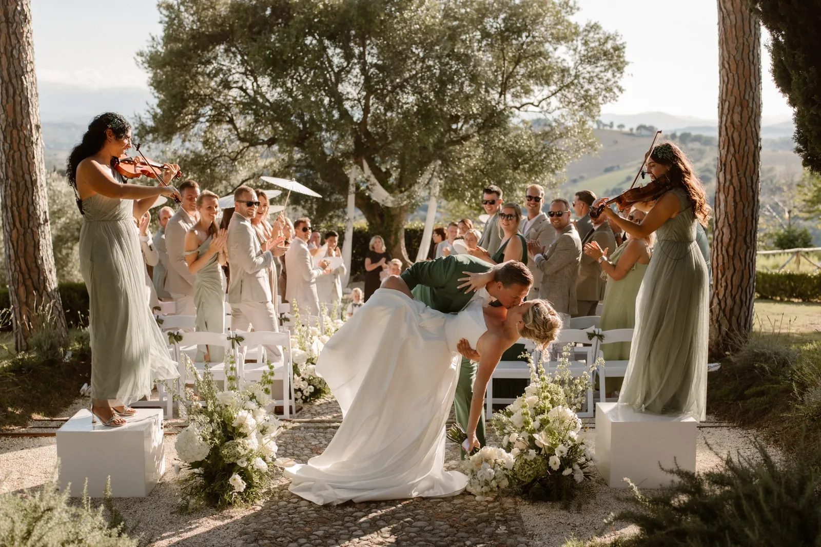 Groom dips the bride for a kiss down the aisle while violinists play under the old olive tree